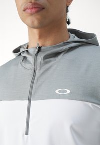 Hoodie gris et blanc avec un design à demi-zip. Présente un tissu texturé et un petit logo blanc sur la poitrine.