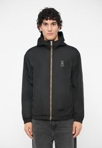 Vyras su garbanotais plaukais, dėvintis juodą švarką su gobtuvu ir užtrauktuku, ant krūtinės turintį "Armani Exchange" logotipą, bei tamsiai pilkas džinsus, prieš baltą foną.