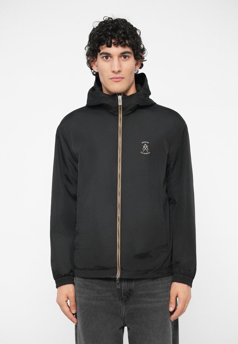 Vyras su garbanotais plaukais, dėvintis juodą švarką su gobtuvu ir užtrauktuku, ant krūtinės turintį "Armani Exchange" logotipą, bei tamsiai pilkas džinsus, prieš baltą foną.