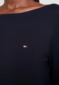 Granatowy sweter z szerokim dekoltem, drobnym splotem i małym haftowanym logo w kolorach czerwonym, białym i niebieskim na piersi.
