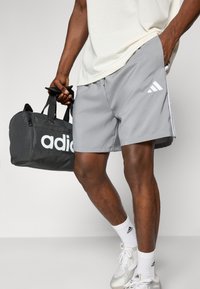 Harmaat urheilushortsit, joissa on valkoiset sivušnaitit ja Adidas-logo; musta laukku, jossa on suuret valkoiset Adidas-brändin logot; valkoiset lenkkarit, joissa on logo.