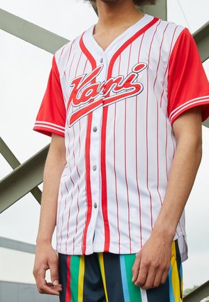 Person iført en hvid og rød Vans baseball-style trøje med lodrette striber og flerfarvede striped bukser, stående nær en metalstruktur.