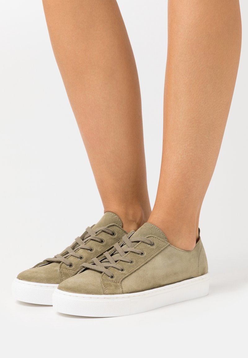 Pieds portant des baskets en daim vert olive à lacets avec semelles en caoutchouc blanc, debout sur une surface blanche.