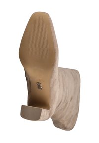 Beige ruskindsankelstøvle med en lav blokhæl, struktureret sål og et lille metalskilt-logo nederst.