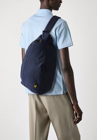 Marineblå slingbag med teksturert stoff, som har en fremre glidelåslomme og en liten gul logo. Båret over skulderen mot en lyseblå skjorte.
