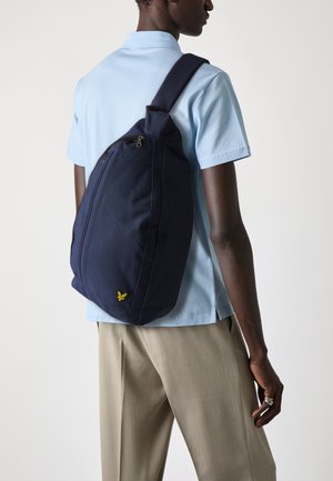 Navy sling bag with a textured fabric, featuring a front zipper pocket and a small yellow logo. Worn over the shoulder against a light blue shirt.