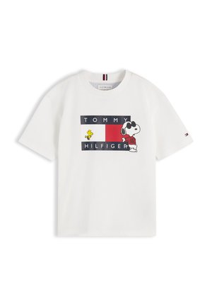 Młoda dziewczyna z dwoma puszystymi kucykami, ubrana w białą koszulkę z grafiką Snoopy’ego, ciemne dżinsy i białe trampki, stoi ze splecionymi rękami.