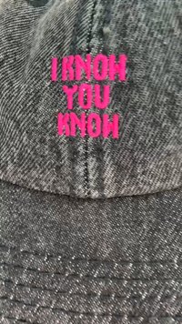 Grådenimkeps med fet, rosa broderad text som står "JAG VET ATT DU VET" på framsidan, med synlig sömnadstextur.