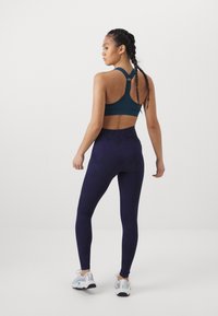 Sportbeha in teal, hoog gewaagde leggings in donkerpaars met subtiel patroon, en witte sportschoenen met blauwe accenten, gezien vanuit de achterkant.