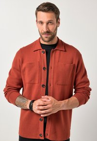 Camicia in pile rossa con bottoni, due tasche frontali, bottoni neri e colletto. Indossata sopra una maglietta nera.