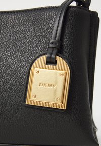 Borsa a mano in pelle nera con superficie testurizzata. Finiture in tonalità oro con un'etichetta rettangolare che mostra "DKNY". Forma arrotondata con cuciture dettagliate.