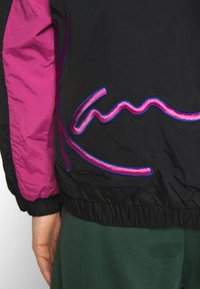 Gros plan d'une manche de veste noire et violette avec un poignet élastique et un dessin brodé abstrait rose et bleu dans le dos.