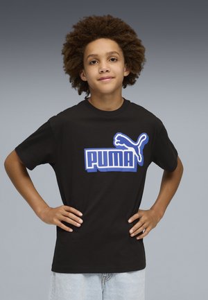 Młoda osoba z kręconymi włosami stoi z rękami na biodrach, ubrana w czarną koszulkę Puma i jasnoniebieskie dżinsy na szarym tle.