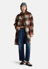 Geruite wollen jas in crème, bruin en oranje. Gecombineerd met donkere jeans en zwarte schoenen. Het model draagt een grijze gebreide beanie.