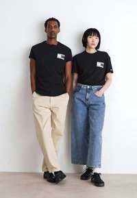 Dos personas llevan camisetas negras con un detalle de bolsillo. Una las combina con pantalones beige, la otra con unos vaqueros de tiro alto azul y zapatillas negras.