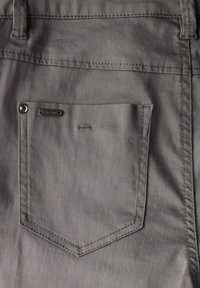 Poche arrière en tissu gris de pantalon avec un coin arrondi, rivet en métal et une petite étiquette noire. Texture lisse et couture minimale.