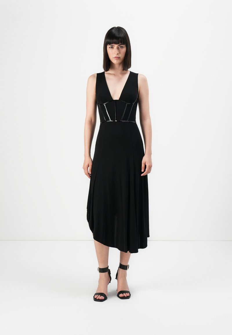 Black dress with a deep V-neck and fitted bodice featuring a glossy accent. Hem drapes asymmetrically. Worn with strappy black heels.