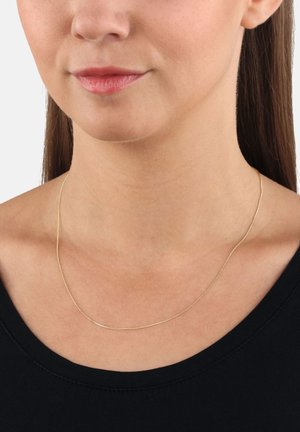 Collana d'oro con un design sottile e liscio, caratterizzata da una forma arrotondata che poggia delicatamente sulla clavicola. Indossata con una maglietta nera semplice.