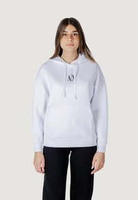Sudadera blanca con un bolsillo frontal, cordones y un pequeño logo negro. Tejido suave con un corte holgado, combinada con pantalones negros.