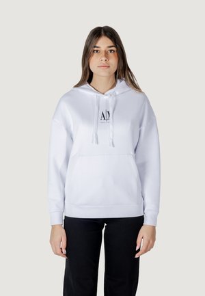 Sudadera blanca con un bolsillo frontal, cordones y un pequeño logo negro. Tejido suave con un corte holgado, combinada con pantalones negros.