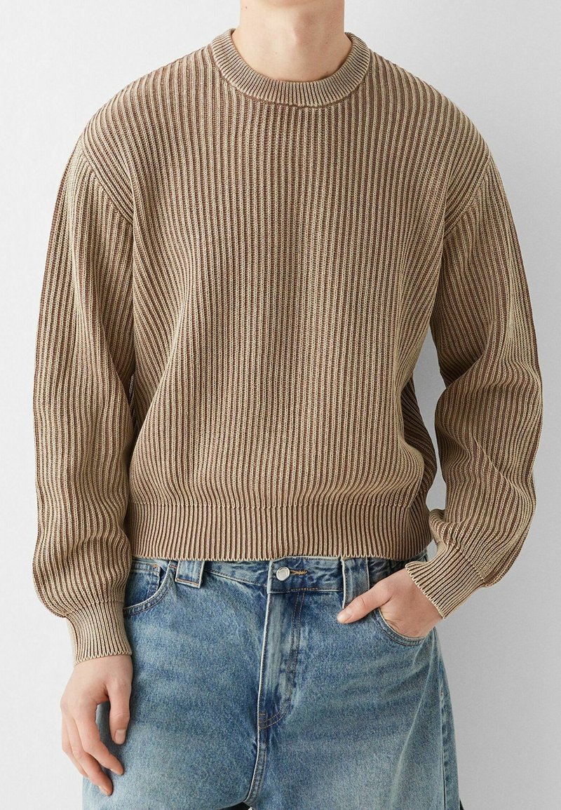 Béžový žebrovaný svetr s kulatým výstřihem a dlouhými rukávy, zdobený texturovanými pruhy a přiléhavým pasem, kombinovaný s džínami s vysokým pasem.
