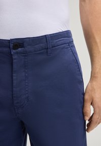 Pantaloni in tessuto testo di colore blu navy, con chiusura a bottone in vita. Presentano una silhouette aderente e dettagli di cucitura minimi.