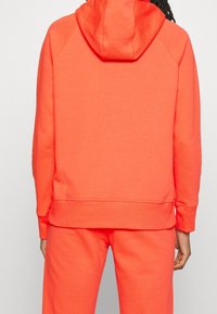 Felpa arancione con una vestibilità rilassata, dotata di cappuccio e polsini a coste, realizzata in tessuto morbido. Il design è semplice, senza ulteriori dettagli.