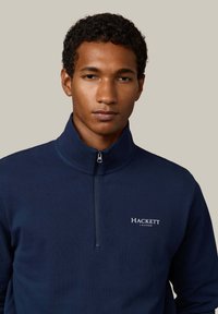 Pullover blu navy con mezza cerniera, realizzato in tessuto morbido; presenta un piccolo logo bianco "HACKETT LONDON" sulla parte sinistra del petto e un motivo testurizzato.