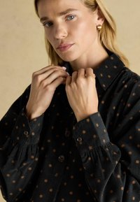 Melnas korduroja krekls ar brūnu zvaigžņu rakstu, ar pogām aizdare, un krokotām piedurknēm; modelis tur audumu pie kakla daļas.