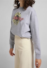 Persona che indossa un felpa grigia con un girasole giallo e la scritta rossa "KANSAS", abbinata a pantaloni crema a vita alta su uno sfondo neutro.