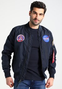 Marineblå bomberjakke med NASA-patches, skinnende nylonmateriale, lynlåslukning, ribbet manchetter og et rødt stofmærke på ærmet.