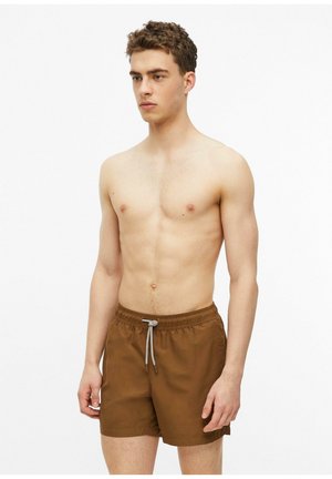 Jeune homme aux cheveux bouclés portant un short de bain marron à cordon, debout avec une expression neutre sur un fond blanc.