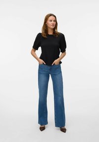 Zwarte textuur top met korte pofmouwen, gecombineerd met blauwe uitlopende jeans. Model staat met de handen in de zakken tegen een effen achtergrond.