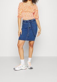Gonna a matita in denim blu con un dettaglio di legatura anteriore, abbinata a un crop top floreale arancione, sneaker bianche e calzini a gambale corto con bordo scallop.
