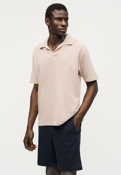 Homme portant un polo beige à manches courtes et un short bleu marine, debout avec une main dans la poche, regardant vers la droite.