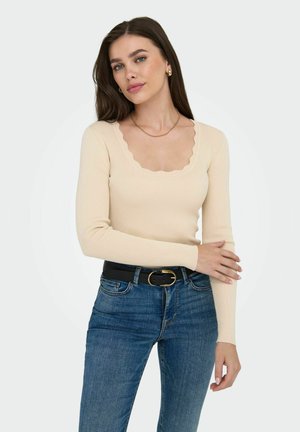 Top de manga larga acanalado en beige claro con escote de pico, combinado con jeans de mezclilla oscura y un cinturón negro con hebilla dorada.