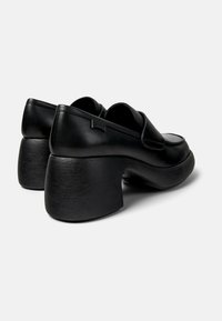 Black leather platform shoes with rounded toes, block heels, and subtle grip texture. Features a Camper logo tag on the heel.