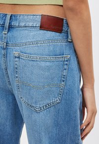 Traperice svijetlo plave boje s smeđim kožnim zakrpama na pojasu i stražnjim džepom koji ima detalje žutog šivanja.