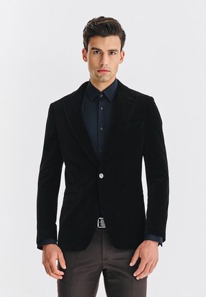 Mężczyzna w czarnym żakiecie na ciemnej koszuli i brązowych spodniach, stojący na tle jednolitego białego tła.