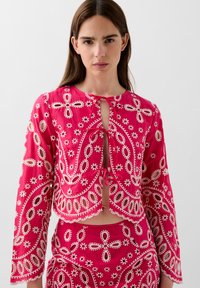 Blouse rose raccourcie avec des motifs floraux brodés, présentant une fermeture à nouer et des manches longues. Le tissu a une texture lisse avec des découpes.