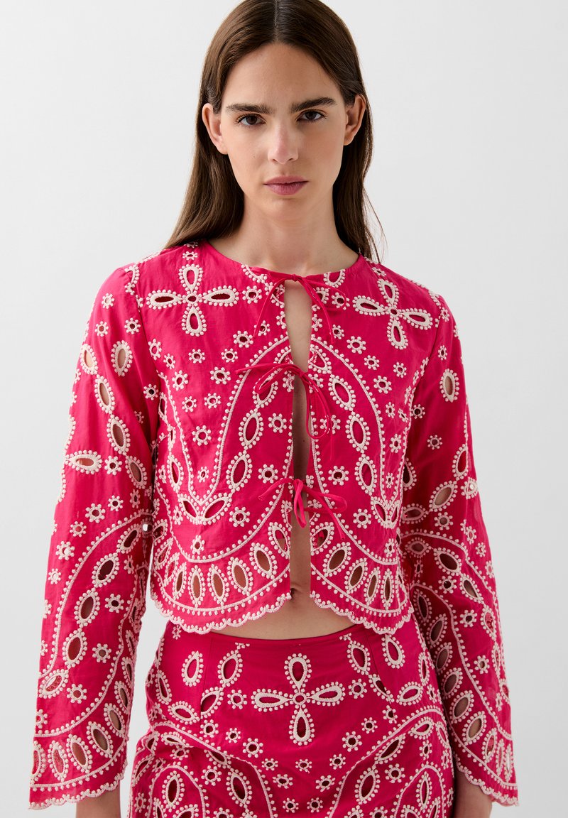 Blouse rose raccourcie avec des motifs floraux brodés, présentant une fermeture à nouer et des manches longues. Le tissu a une texture lisse avec des découpes.