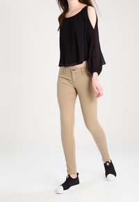 Svart plisserad crop top med kalla axlar, beige skräddarsydda byxor och svarta slip-on sneakers med vita sulor. Enkel, avslappnad design genomgående.