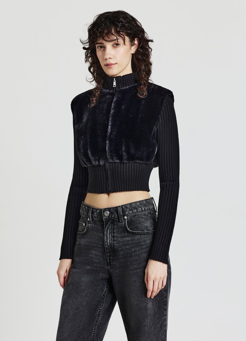 Schwarze, verkürzte Weste mit Vorderseite aus Kunstfell und gerippten langen Ärmeln; hoher Kragen und Frontreißverschluss. Kombiniere sie mit hochtaillierten schwarzen Jeans.