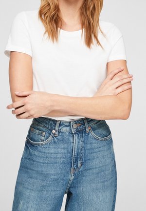 Person iført en simpel hvid kortærmet skjorte og højtaljede blå denimjeans med armene krydset over brystet.