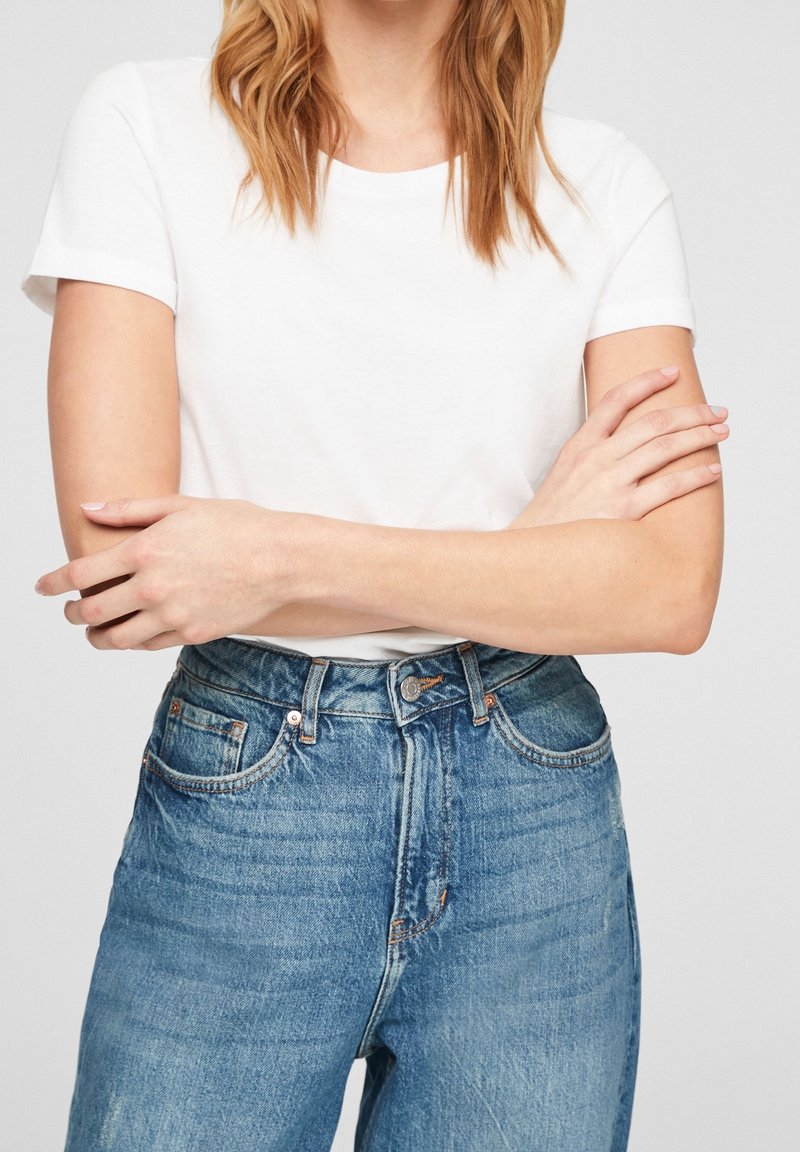 Personne portant un t-shirt blanc uni à manches courtes et un jean bleu taille haute, bras croisés sur la poitrine.