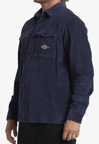 Navy corduroy shirt with vertical stripes, button-up front, two chest pockets, and an embroidered logo on the left pocket.