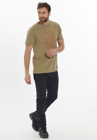 Olivgrön t-shirt med en bergsgrafik och text i orange, kombinerad med svarta byxor och robusta svarta vandringsskor.