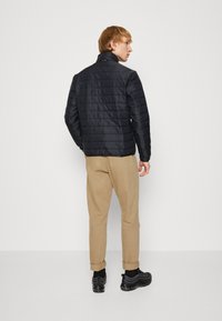 Zwarte gewatteerde jas met een hoge kraag, met een textuurafwerking en een verticale gestikte design, gedragen met beige broeken en zwarte sneakers.