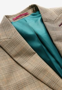 Gros plan d'un blazer marron à carreaux avec de larges revers et une doublure intérieure lisse bleu sarcelle visible.