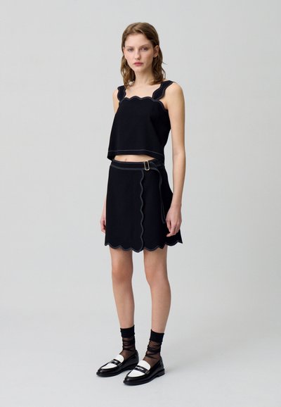 Haut court noir à bords festonnés avec des coutures contrastantes, associé à une jupe noire à bords festonnés ornée d'un détail de boucle sur le côté. Le mannequin porte des chaussures noir et blanc.